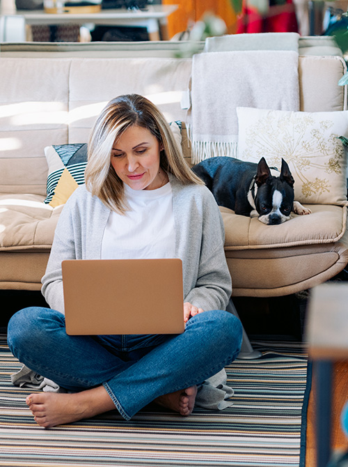 Woman sitting on living room floor using laptop next to sofa where her dog is resting